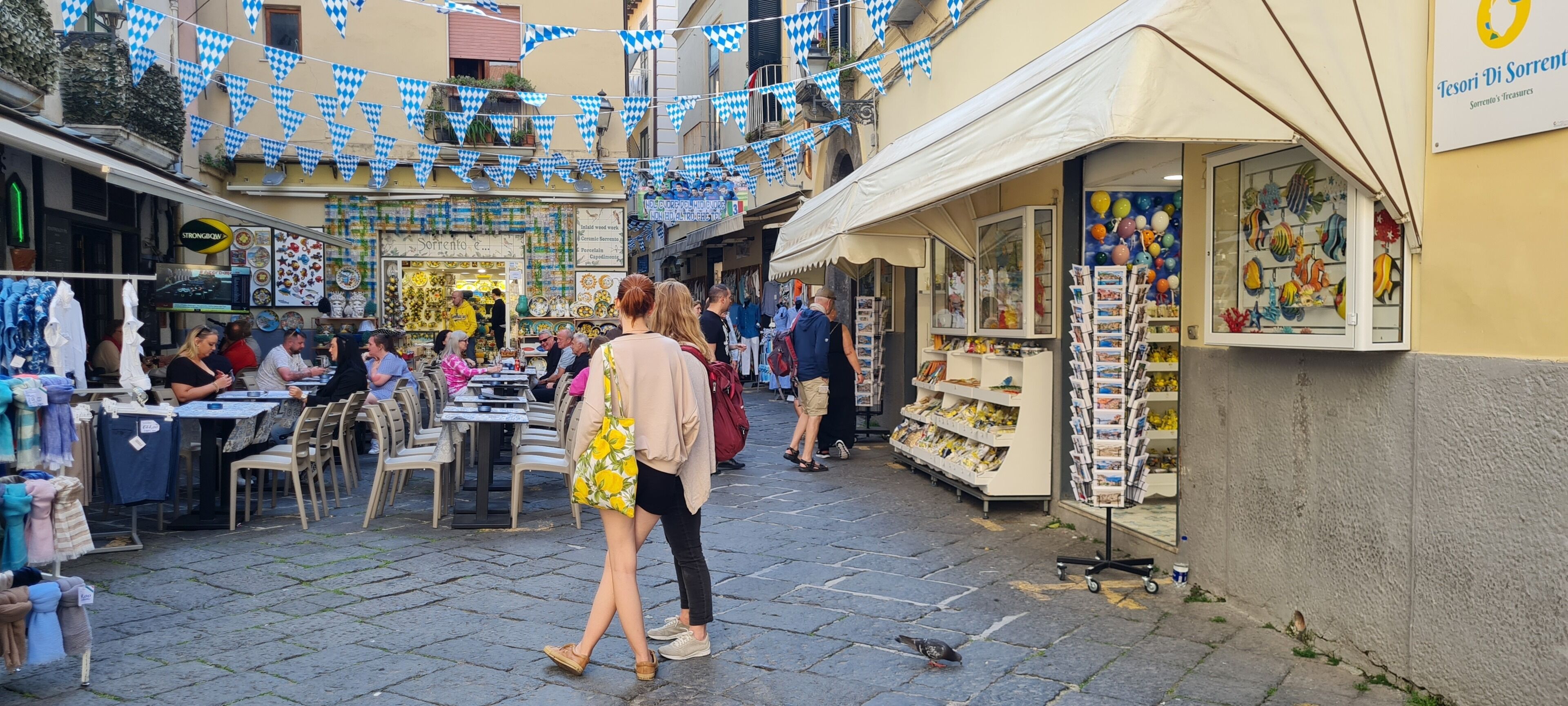 Rua com mesas ao ar livre, lojas e um letreiro que diz 'Tesori Di Sorrento'.