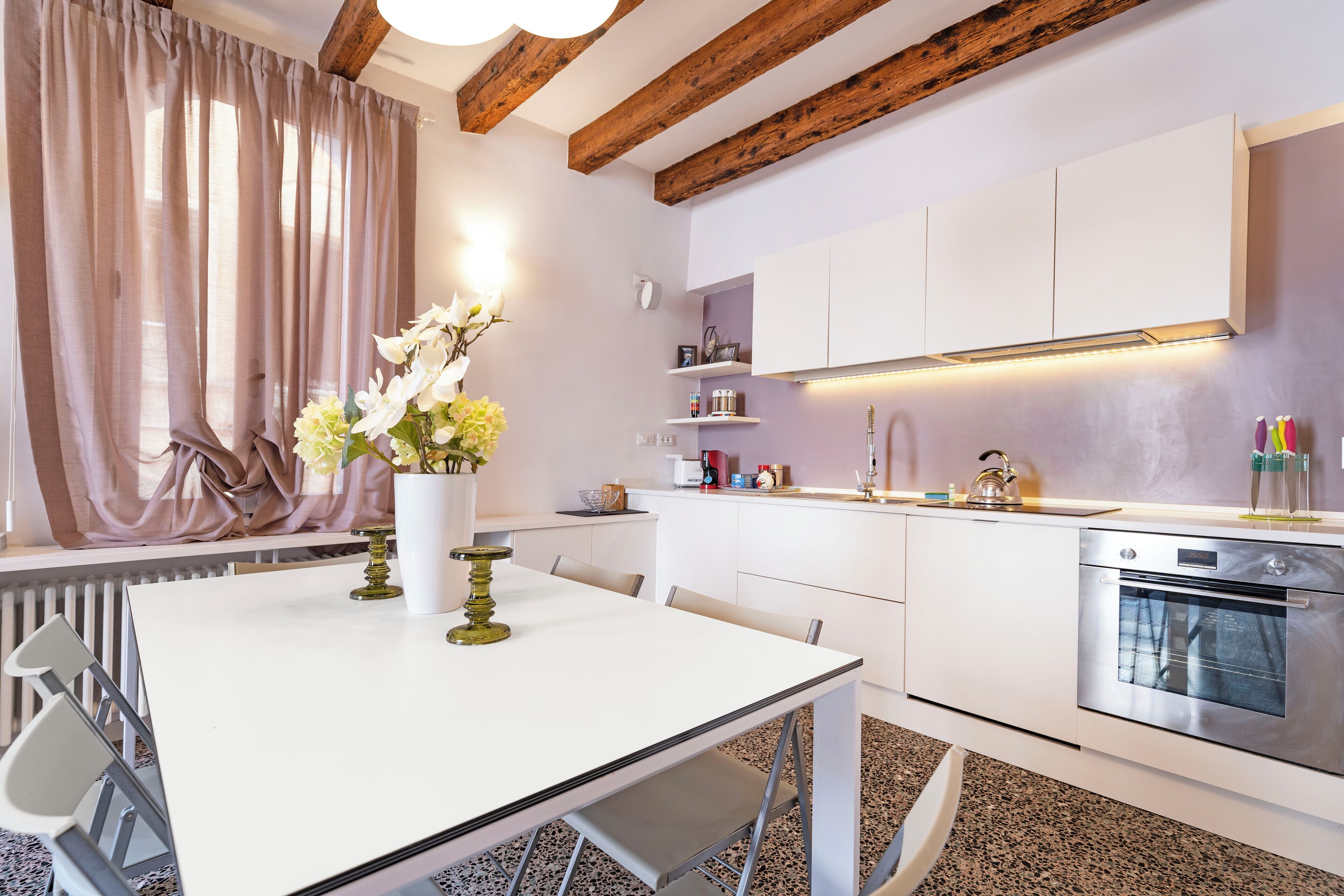 A modern kitchen with white cabinetry, a stainless steel oven, and a dining area with a white table and chairs.