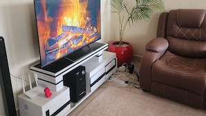 Un salon doté d’un téléviseur à écran plat qui diffuse une image de cheminée, d’un canapé en cuir marron, d’une plante en pot et d’un meuble de rangement blanc agrémenté d’un objet décoratif rouge.
