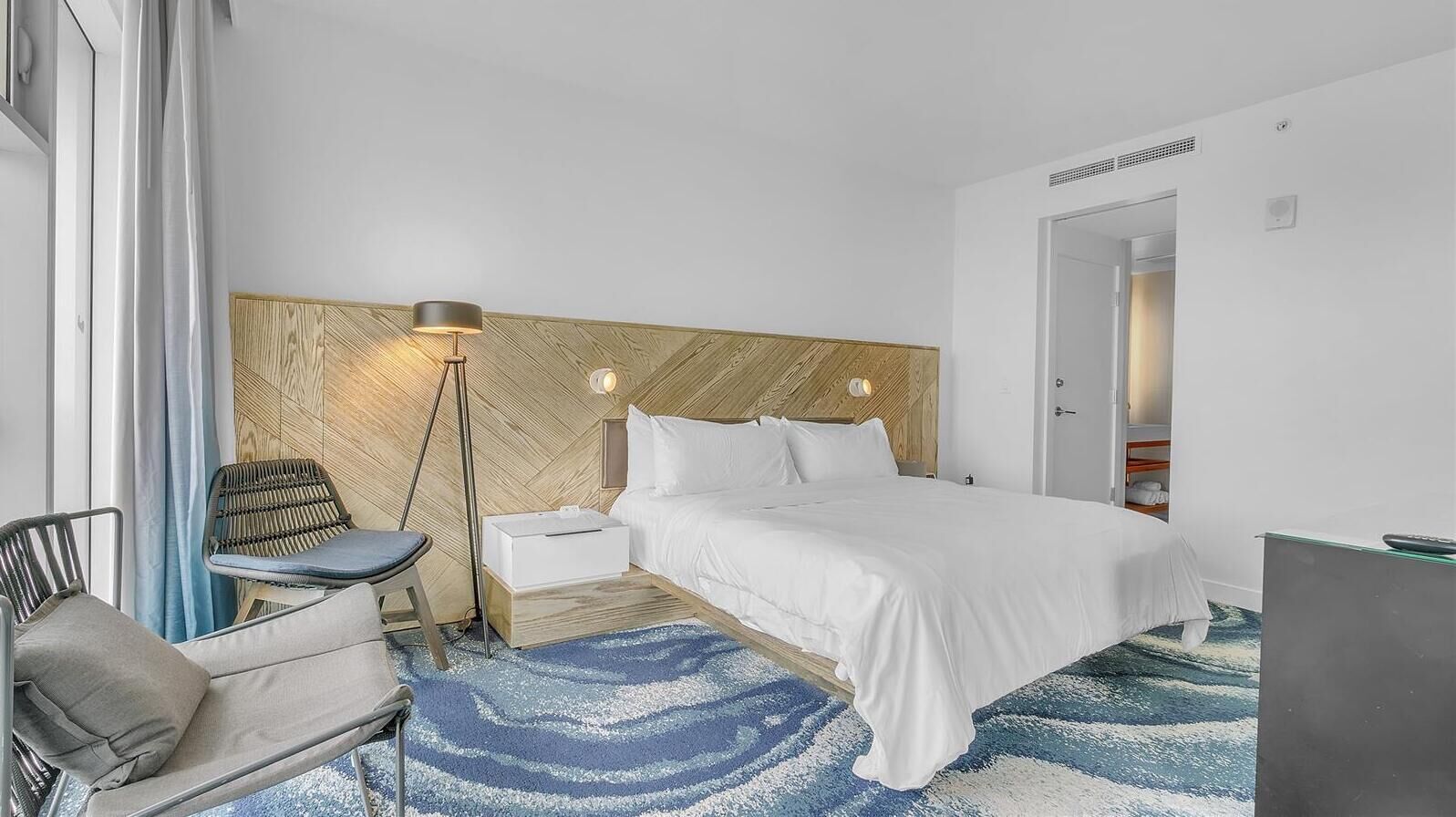 A modern hotel room with a large bed, a bedside table, a lamp, a chair, and a blue patterned rug.