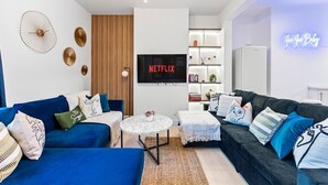 Ein modernes Wohnzimmer mit einem blauen Sofa, einem schwarzen Sofa, einem weißen Couchtisch und einem an der Wand befestigten Fernseher, der 'NETFLIX' anzeigt.