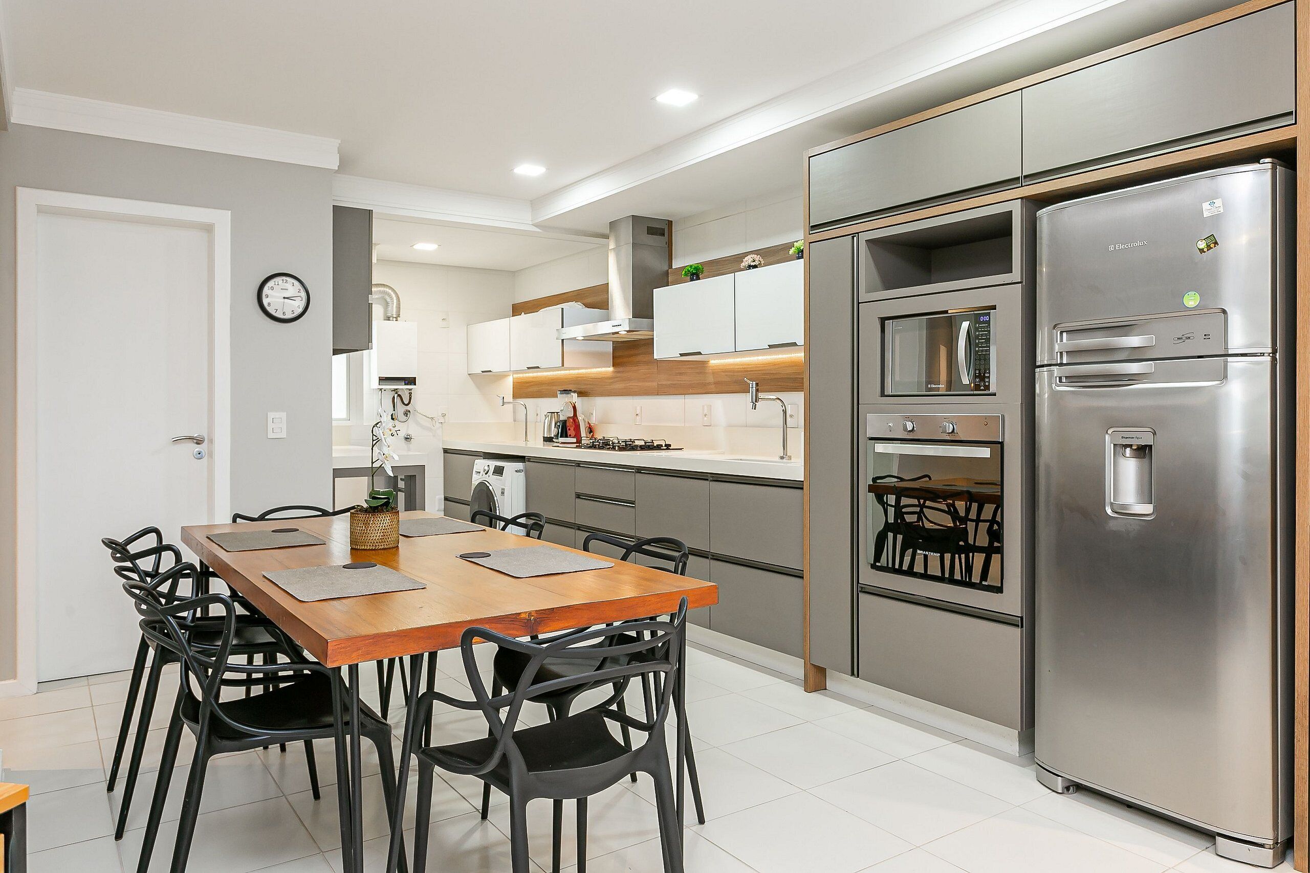 A modern kitchen with a wooden dining table, black chairs, stainless steel appliances, and a clock on the wall.