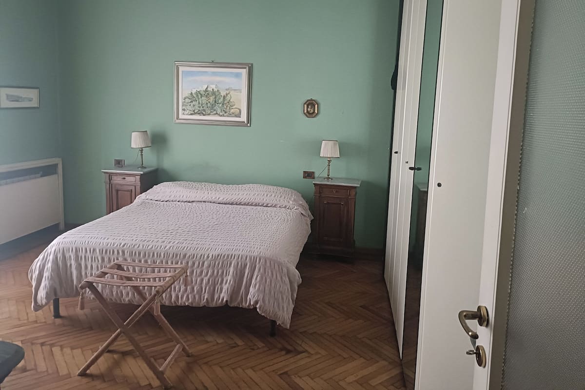 A bedroom with a bed, two bedside tables with lamps, a wooden floor, a door, and a framed picture on the wall.