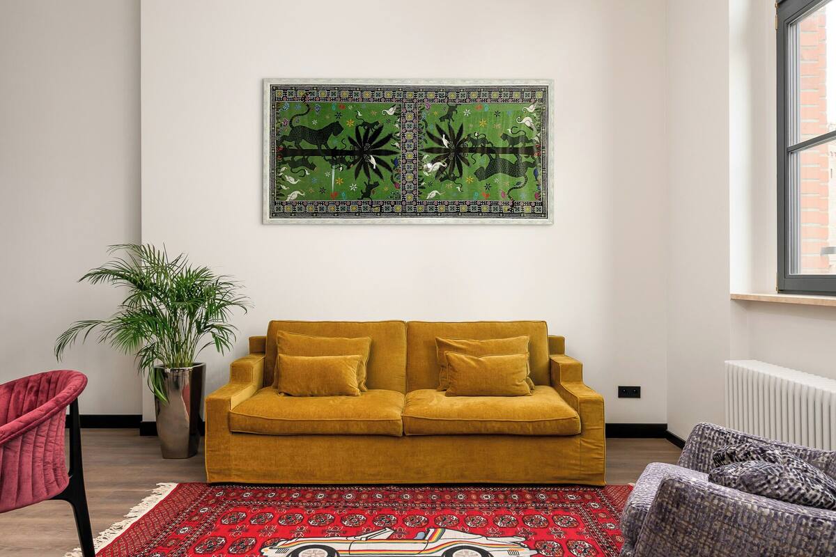 Un salon avec un canapé moutarde, un tapis à motifs rouges, une plante en pot et un tableau abstrait accroché au mur.