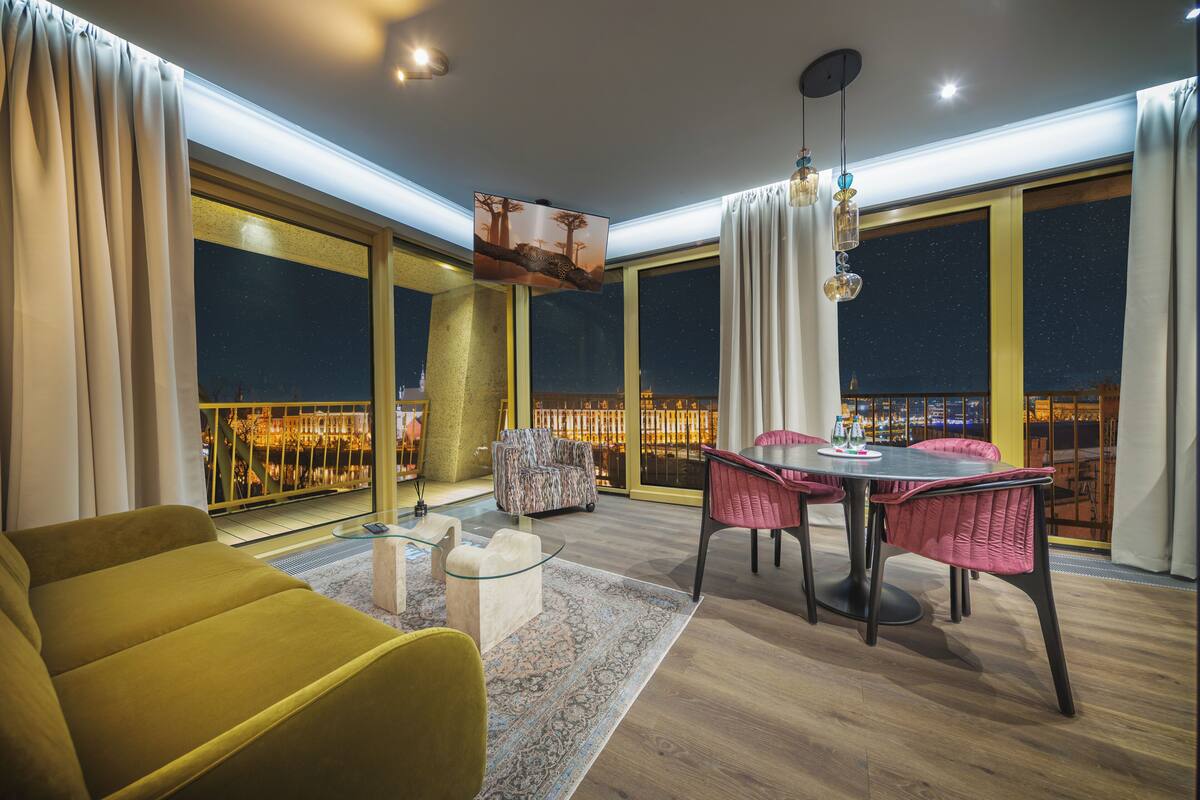 Un salon moderne comprenant un canapé, une table à manger avec des chaises, un téléviseur et de grandes fenêtres offrant une vue sur le paysage urbain la nuit.