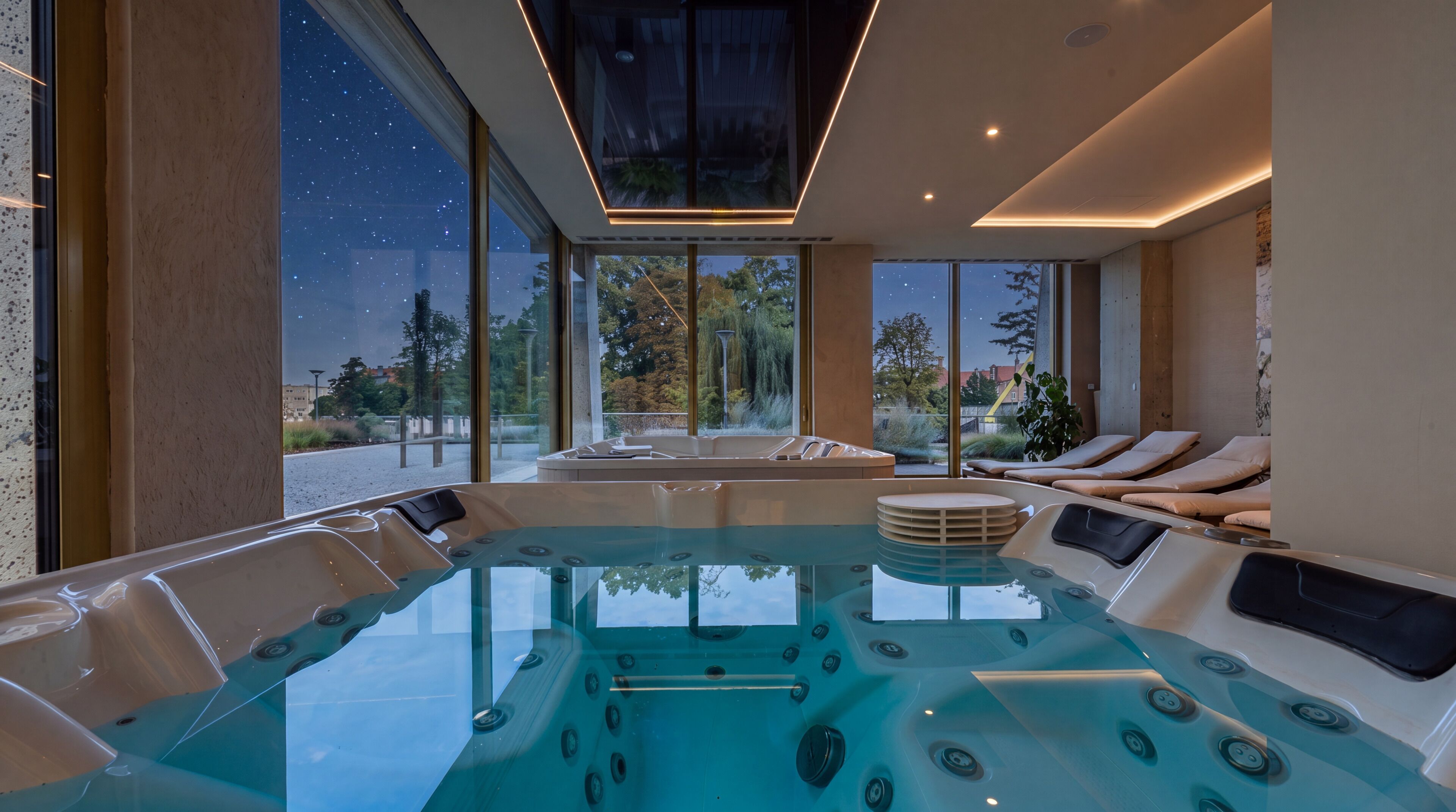 Una piscina interior moderna con cerramiento de vidrio, rodeada de sillones de descanso y vistas al exterior.