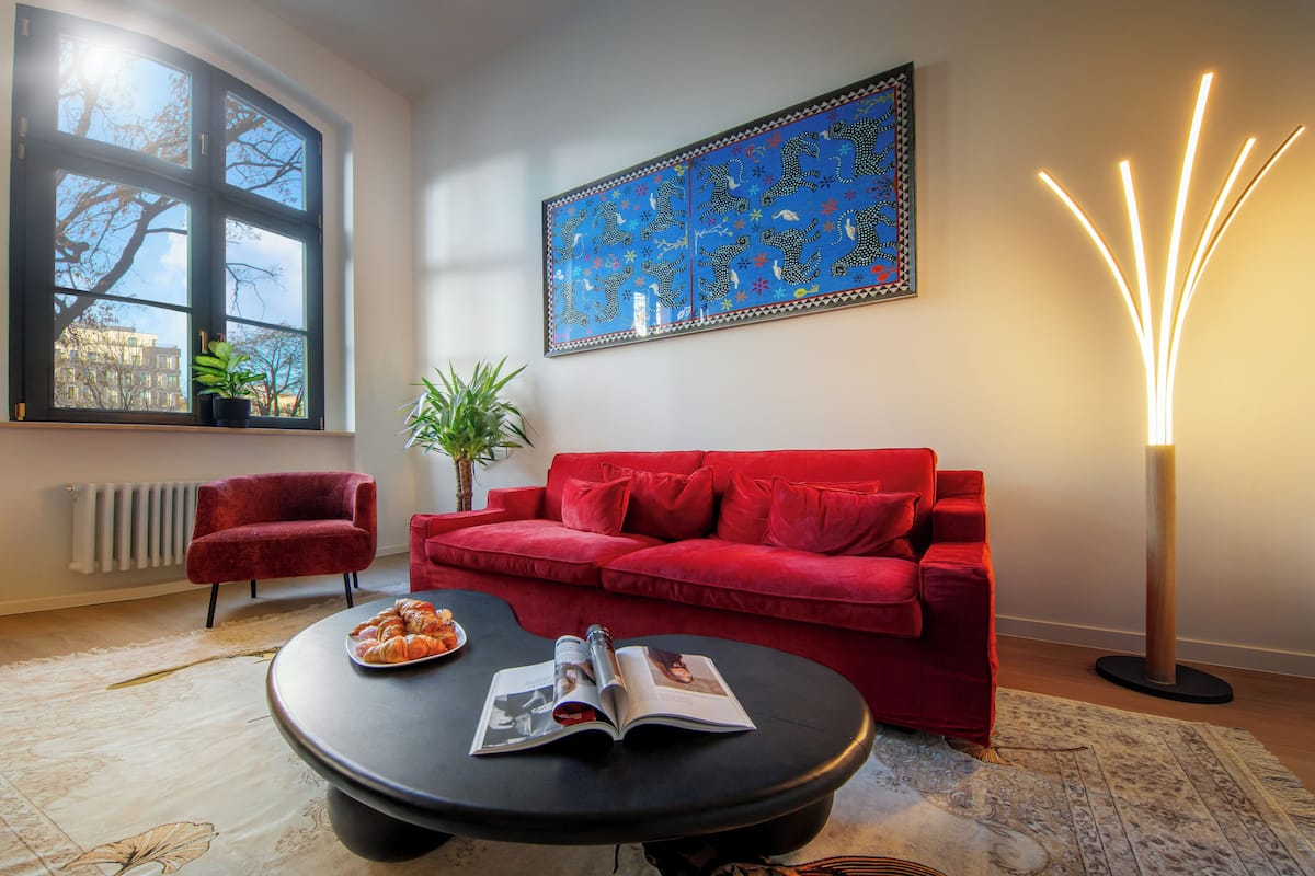 Un salon avec un canapé rouge, une table basse noire sur laquelle se trouvent un magazine et un croissant, une grande fenêtre et un lampadaire.