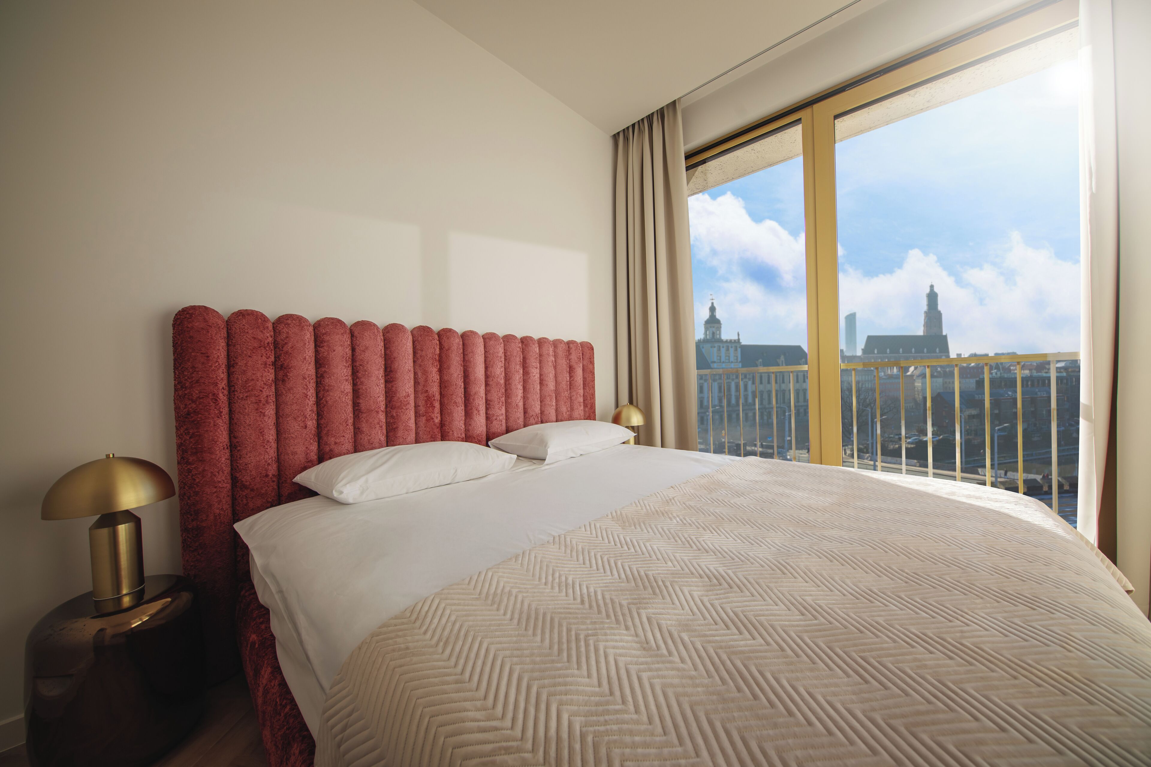 Hotelkamer met een groot bed, nachtkastje met een lamp en uitzicht op een stadsgezicht door een groot raam.