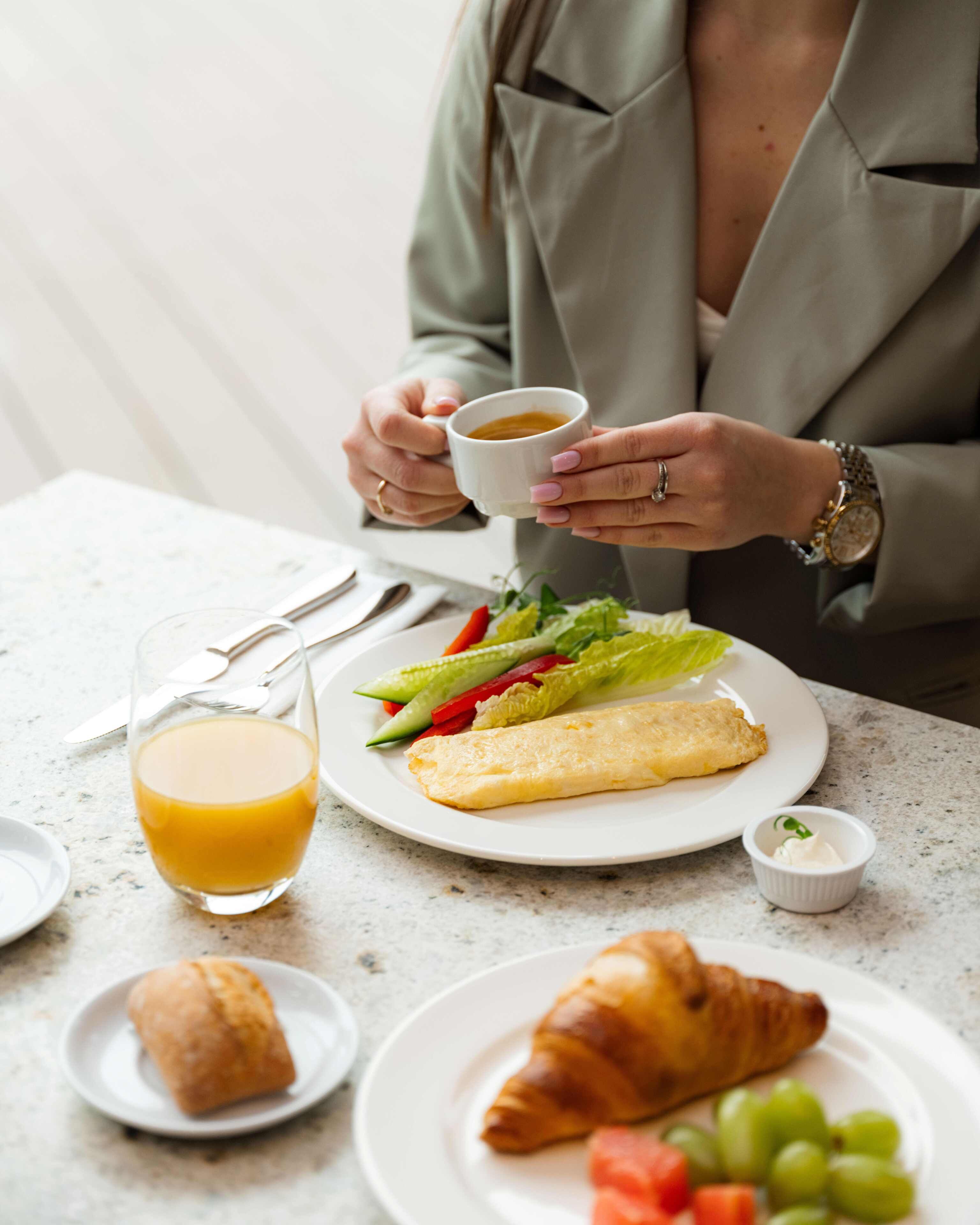 Eine Person hält eine Tasse, sitzt an einem Tisch mit Tellern voller Essen, einem Glas Orangensaft und einem Croissant.
