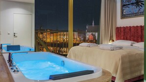 A hotel room with a large window, a bed, a bathtub, and a view of a cityscape at night. - Boulevard Drobnera Residence (Wroclaw)