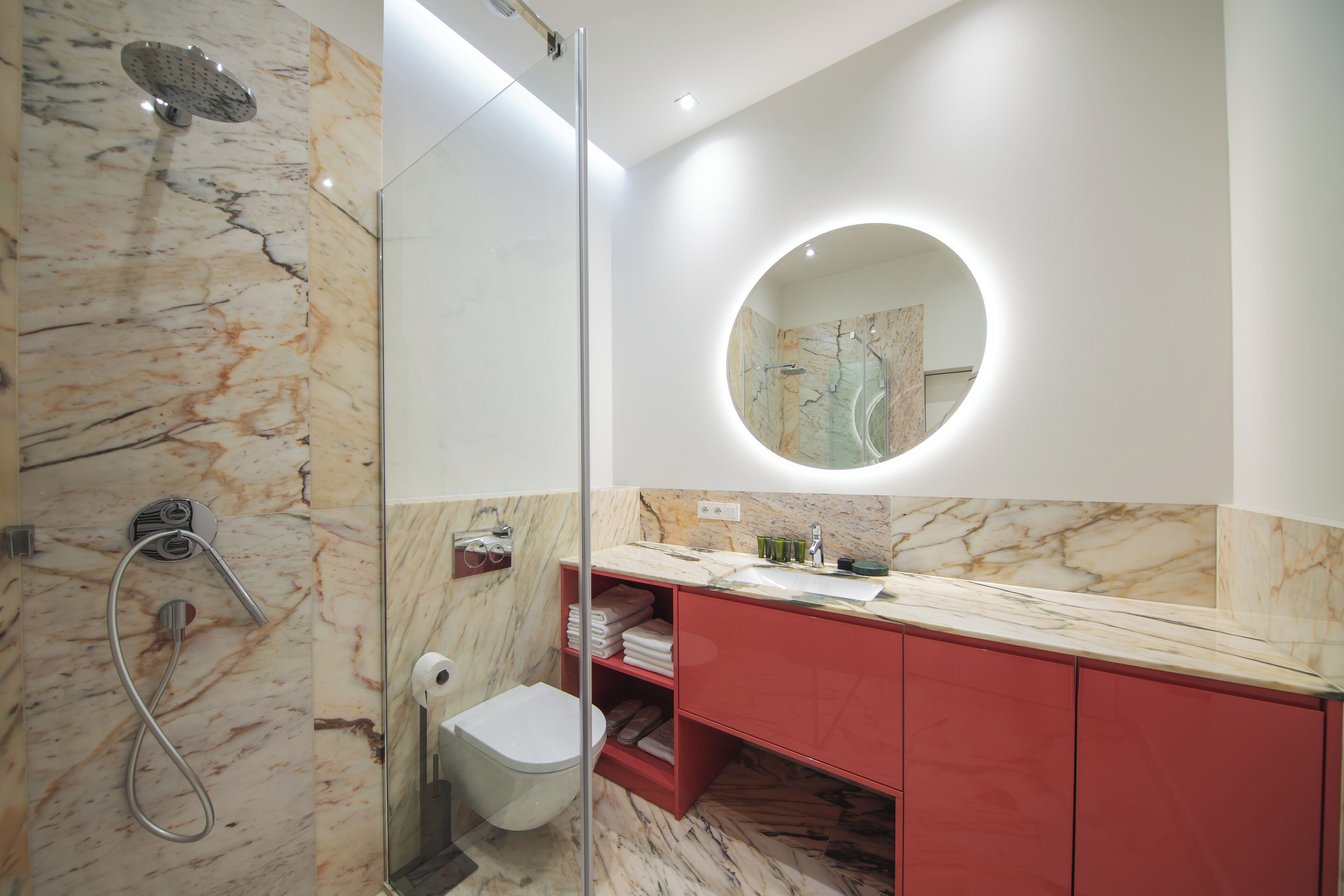 Un bagno moderno con doccia in marmo, servizi igienici racchiusi in vetro, mobile lavabo rosso e specchio tondo.