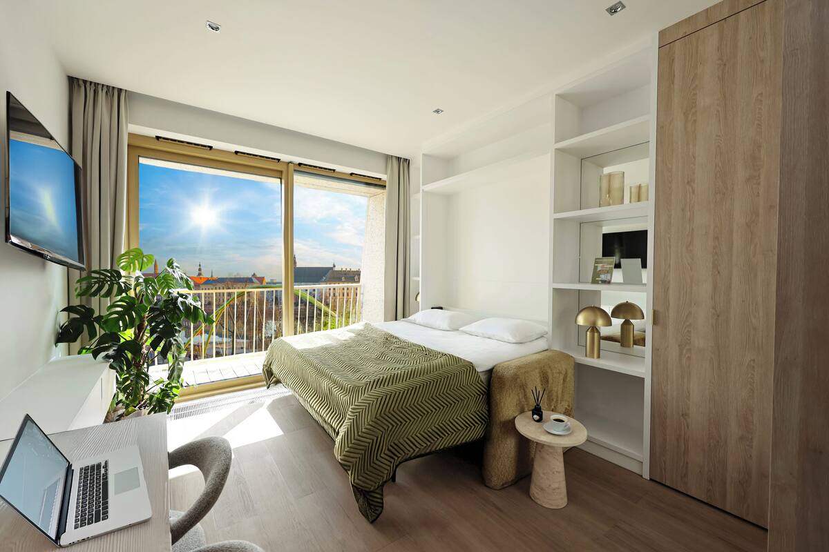 Une chambre d’hôtel moderne équipée d’un lit, d’un bureau avec un ordinateur portable, d’une petite table avec une lampe et d’un balcon avec vue.