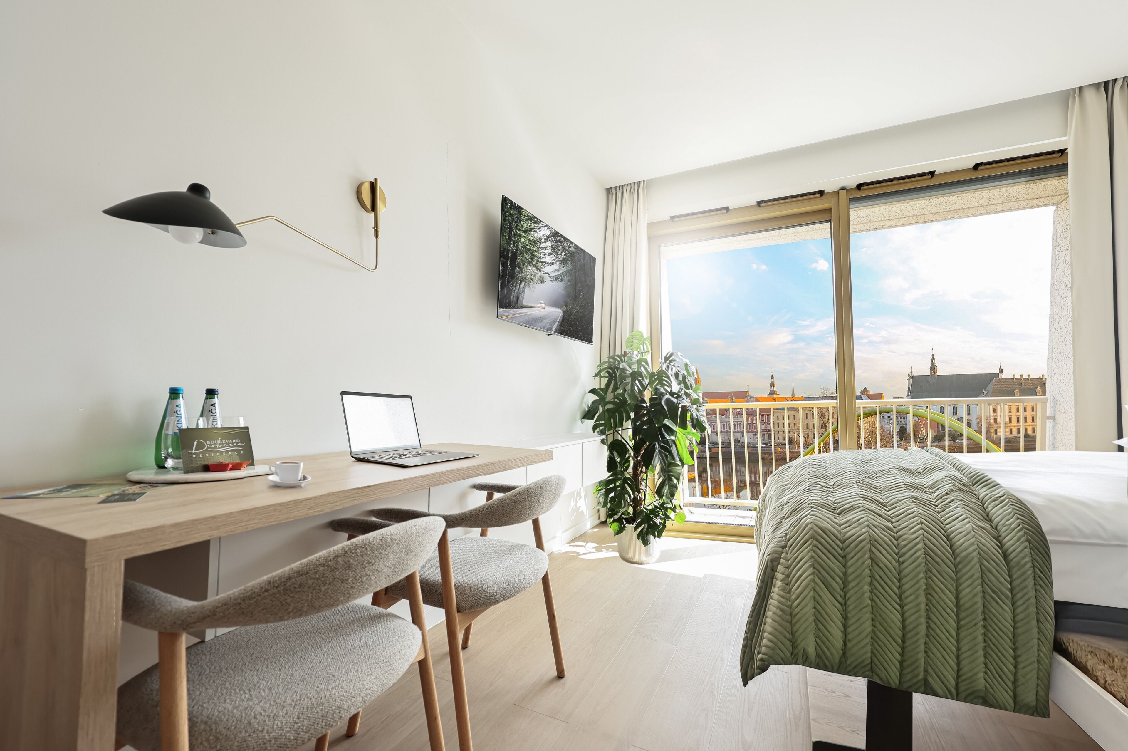 Une chambre d’hôtel moderne avec un bureau en bois, un ordinateur portable et deux chaises. Une grande fenêtre offre une vue sur le paysage urbain.