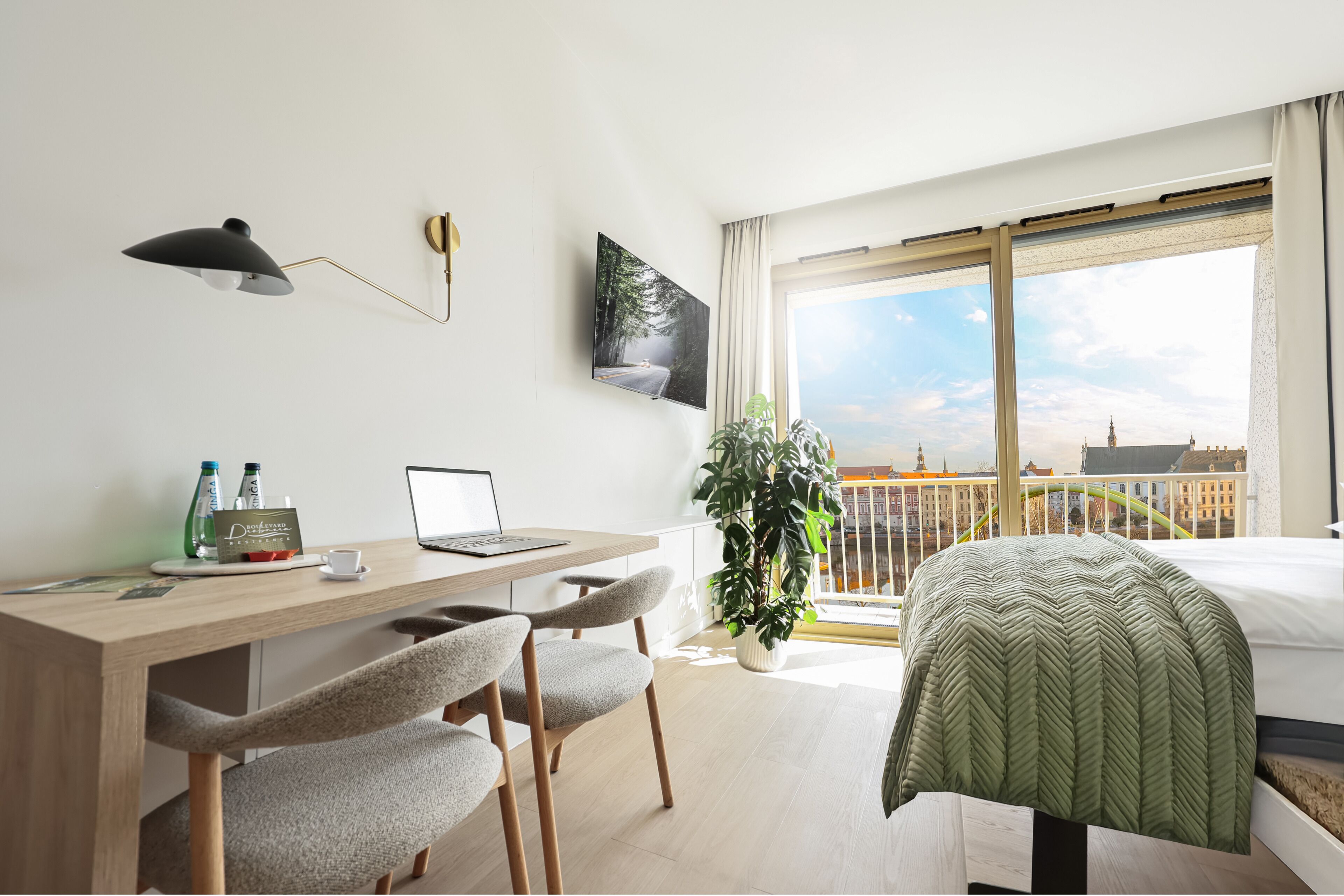 A modern hotel room with a wooden desk, laptop, and two chairs. A large window offers a view of a cityscape.