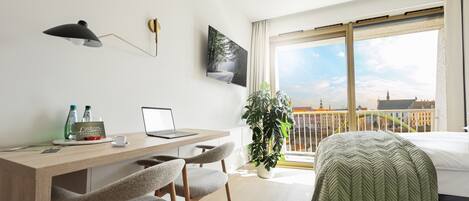 A modern hotel room with a wooden desk, laptop, and two chairs. A large window offers a view of a cityscape.