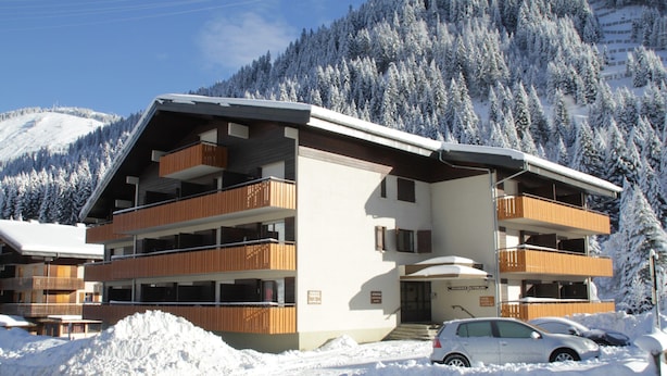 Een meerhoog hotel met balkons, een piste op de achtergrond en een met sneeuw bedekte parkeerplaats.