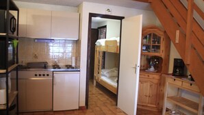 Een compacte keuken met een ingebouwde kookplaat, een vaatwasser en een houten kastje. Een stapelbed is zichtbaar door een open deur in de aangrenzende kamer.