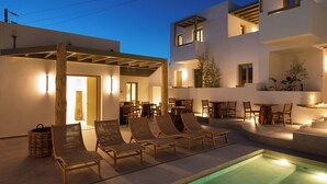A modern outdoor pool area with lounge chairs and tables, surrounded by a white building with balconies. - Mazee Paros (Paros)