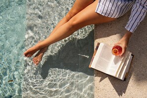 A person sitting by a poolside with a book and a drink.