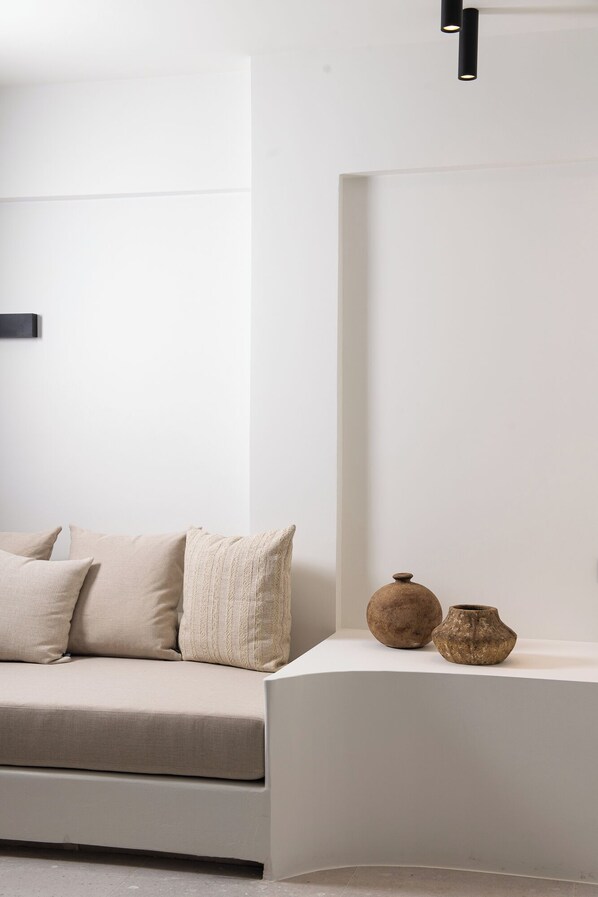 A minimalist living room with a sofa, cushions, and two pottery pieces on a white console table. - Mazee Paros (Paros)