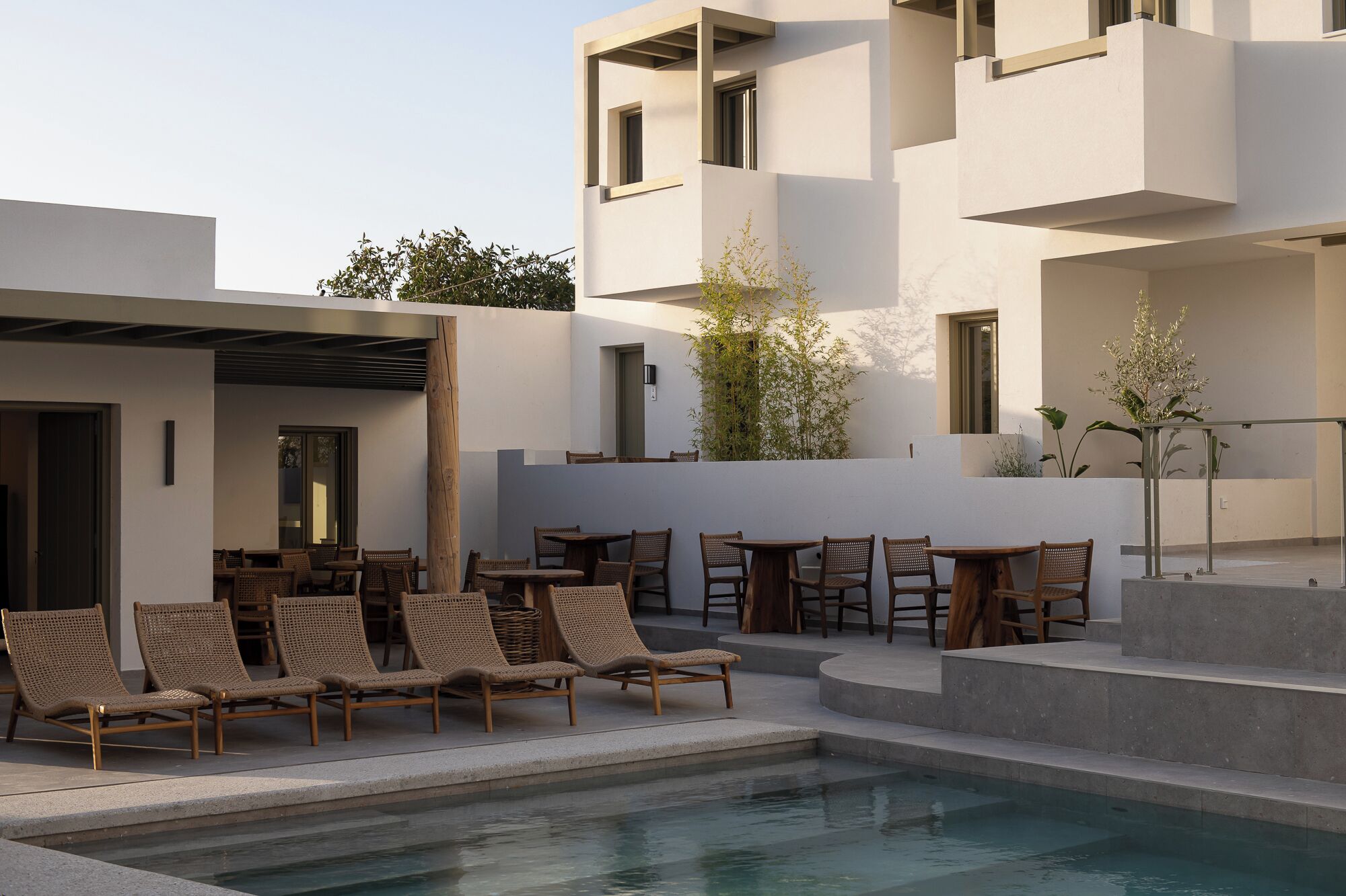 A modern outdoor pool area with lounge chairs and tables, surrounded by a minimalist building with balconies.