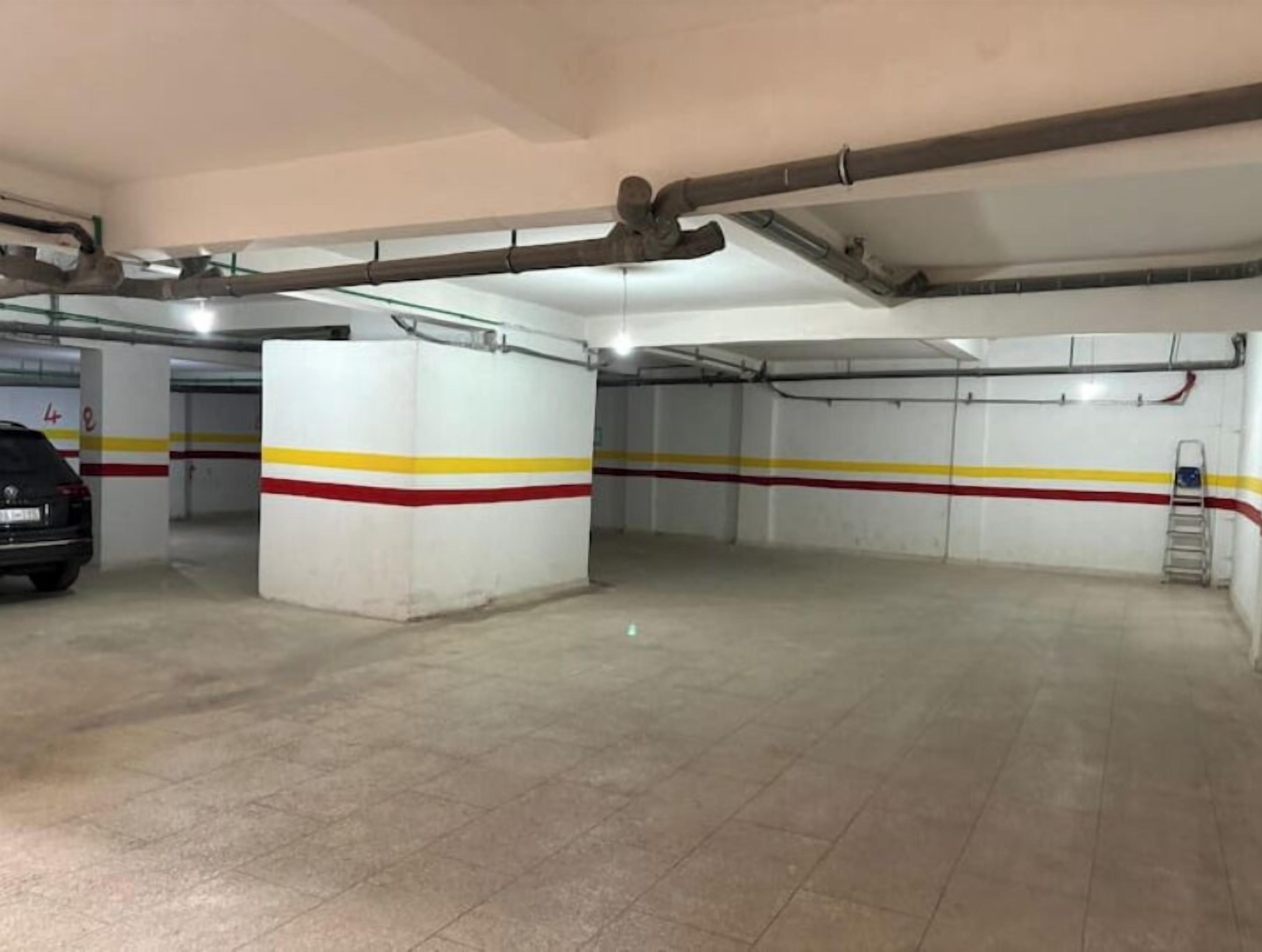Un estacionamiento con paredes blancas, franjas amarillas y rojas, y un coche estacionado en una de las plazas.