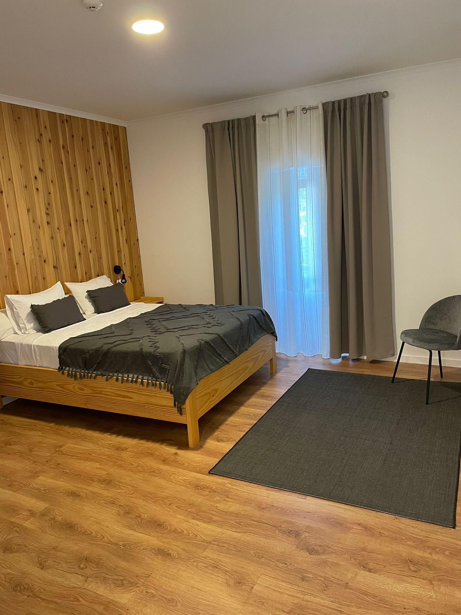 Una camera da letto moderna con un letto di legno, una poltrona grigia e un'ampia finestra con tende.