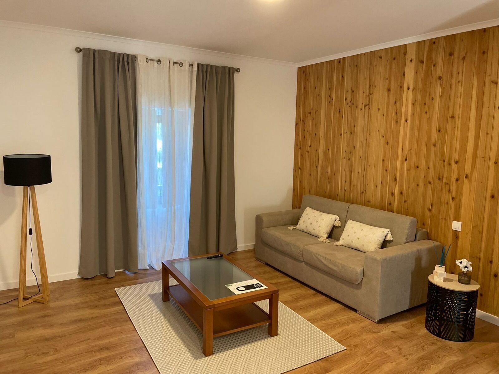 Een woonkamer met een houten vloer, een bank, een salontafel met glazen blad, een lamp en een houten wandpaneel.