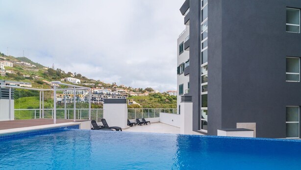 Une piscine sur le toit, agrémentée de chaises longues, offrant une vue sur une colline parsemée de bâtiments, et un immeuble moderne doté de grandes fenêtres.