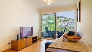 Un salon moderne doté d’un téléviseur à écran plat, de meubles en bois, d’un balcon avec des chaises et offrant une vue sur une colline.