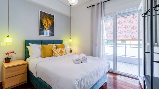 Un lit bien fait, avec du linge de lit blanc, une tête de lit verte et des coussins jaunes et blancs. On aperçoit une petite table de chevet avec une lampe et une plante en pot. Un tableau est accroché au-dessus du lit, et une fenêtre avec des rideaux est visible.