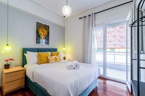 A neatly made bed with white linens, a green headboard, and yellow and white pillows. There is a small bedside table with a lamp and a potted plant. A painting hangs above the bed, and a window with curtains is visible. - Psirri Urban Chic · Central 3BR · Sleeps 10 (Athina)