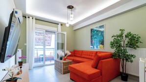 Un salon moderne avec un canapé d’angle, un téléviseur à écran plat, un luminaire au plafond et une grande fenêtre donnant sur l’extérieur.