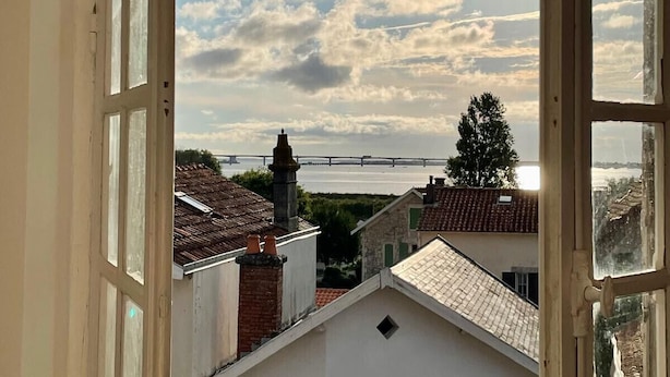 Une vue à travers une fenêtre ouverte donnant sur un balcon, un pont au loin et des bâtiments aux toits couverts de tuiles.