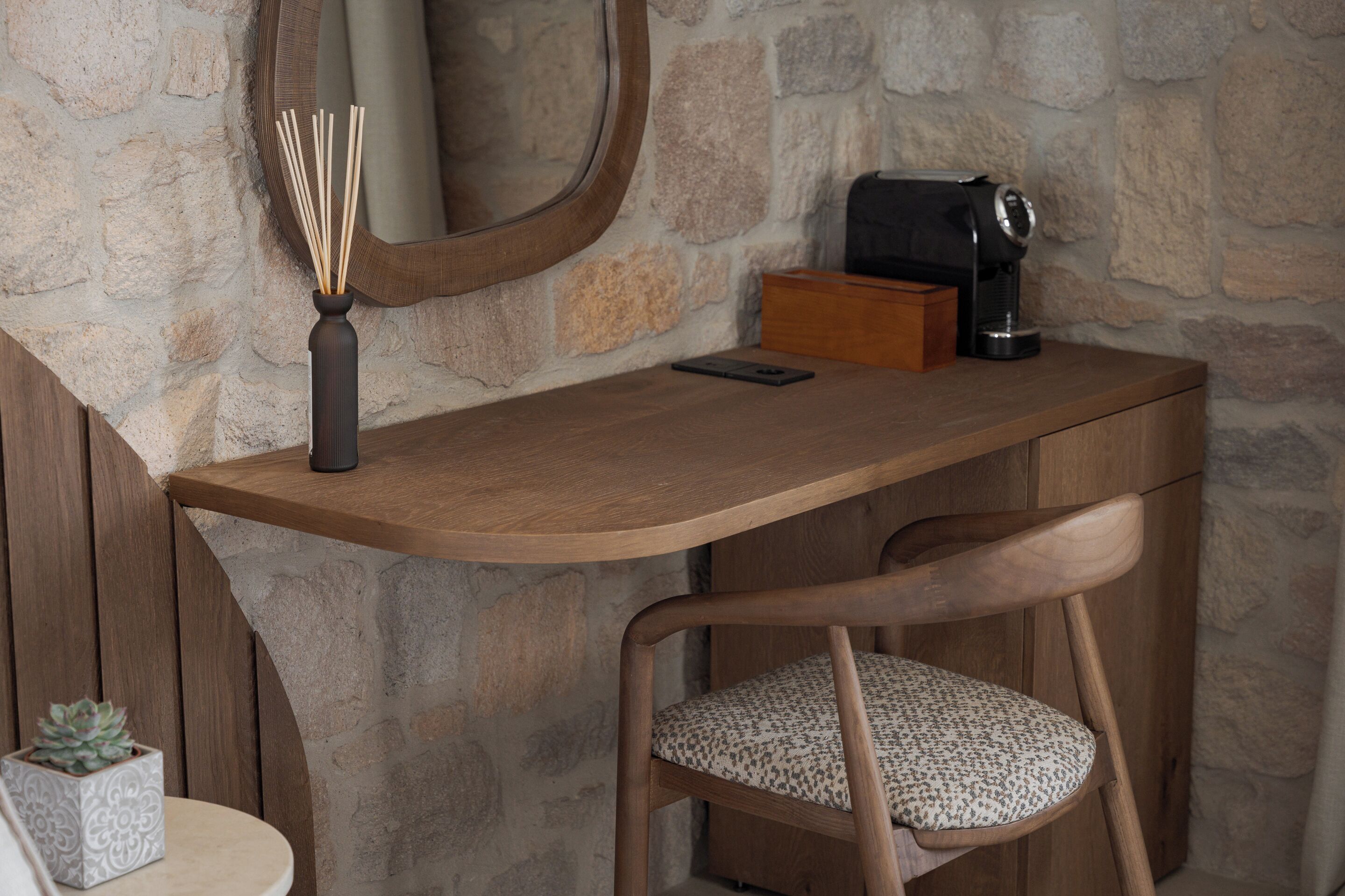 A wooden desk with a mirror, a vase with reeds, a coffee maker, and a chair with a patterned cushion.