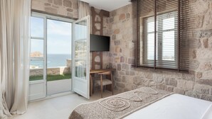 Een slaapkamer met een stenen muur, een bed, een bureau, een stoel, een televisie en uitzicht op zee.