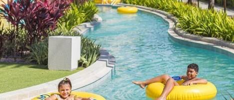 A pool area with two children on yellow inflatable tubes, surrounded by palm trees and a water slide.