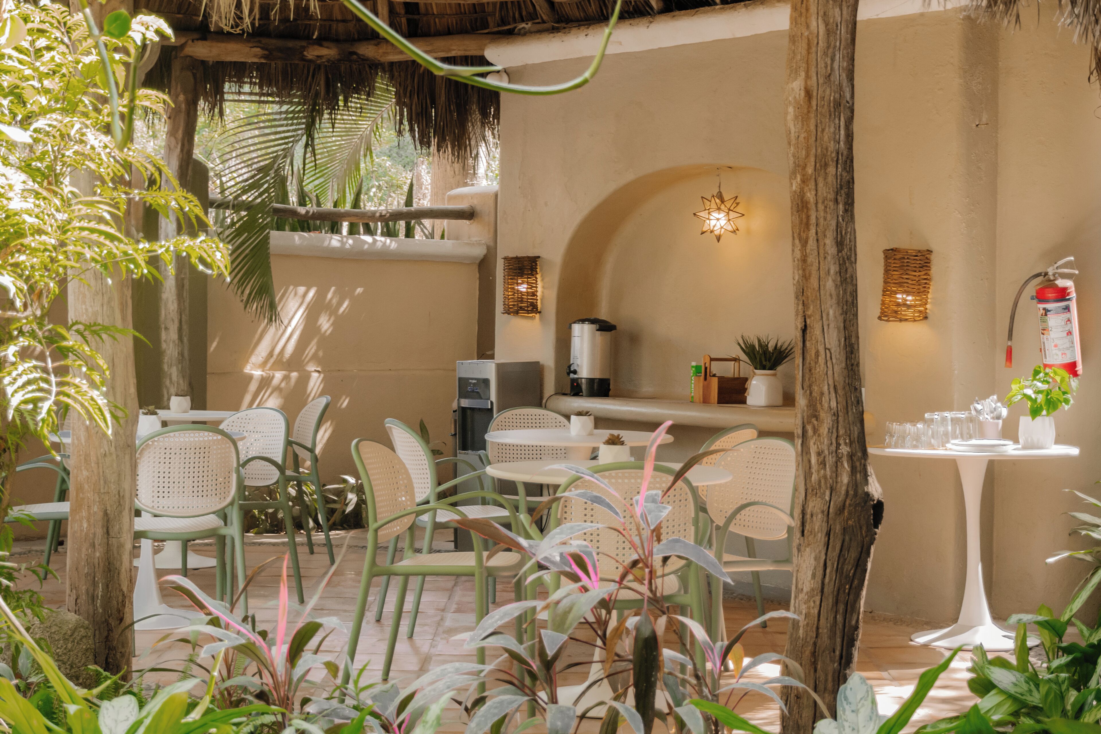 An outdoor seating area with green chairs and tables, surrounded by plants and a thatched roof.