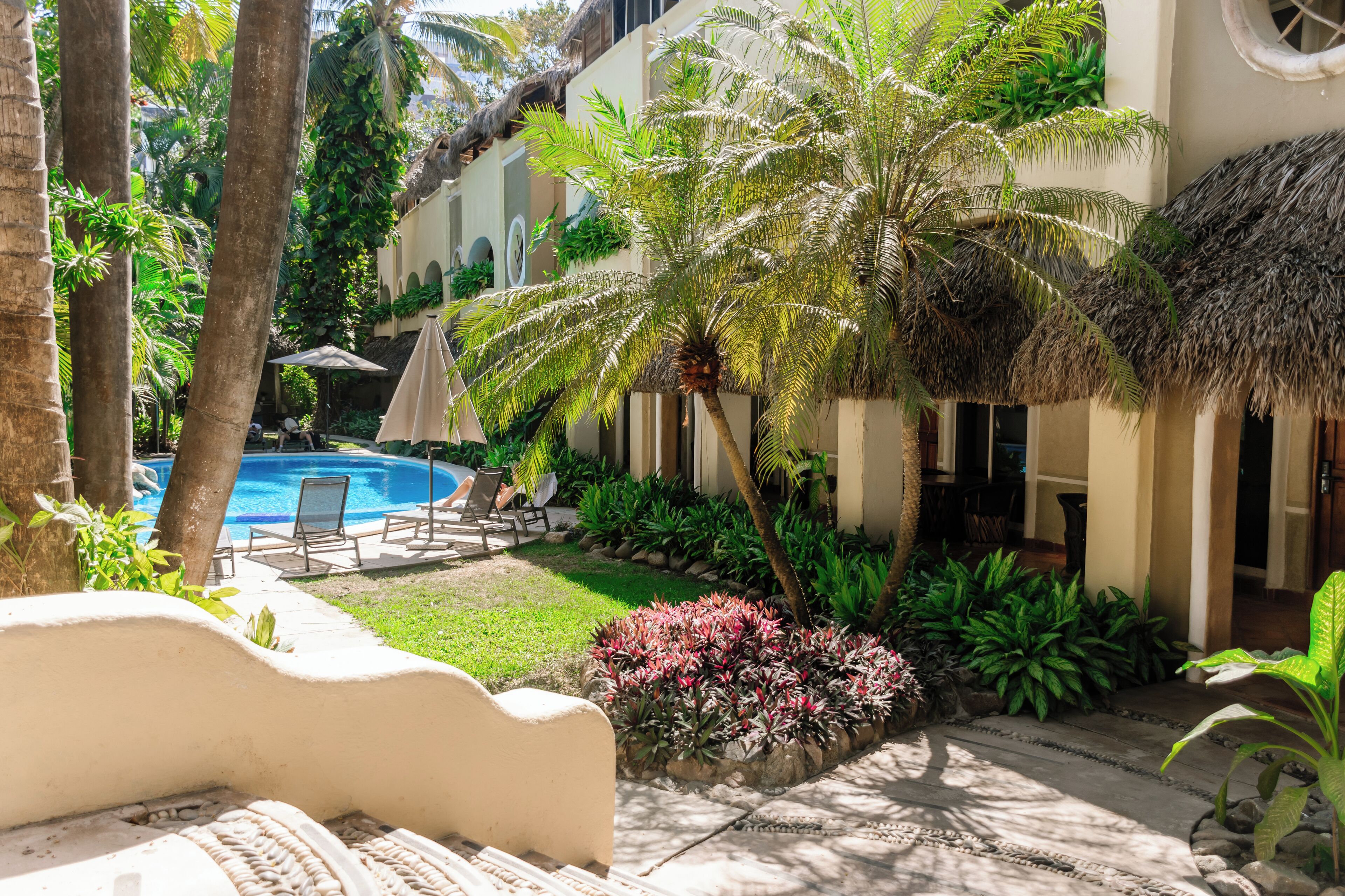 Zona de piscina con sillones de descanso, palmeras y una estructura con techo de paja.