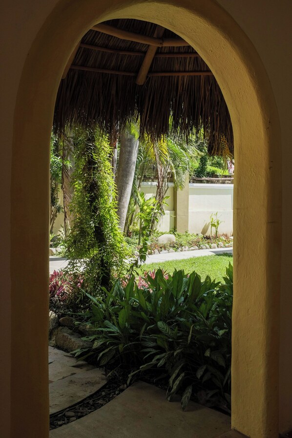 Un arco d'ingresso conduce a un giardino con piante tropicali e una struttura con tetto di paglia.