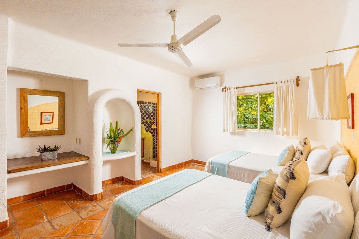 Une chambre d’hôtel avec deux lits, un ventilateur de plafond, un petit bureau, une fenêtre avec des rideaux et une plante.