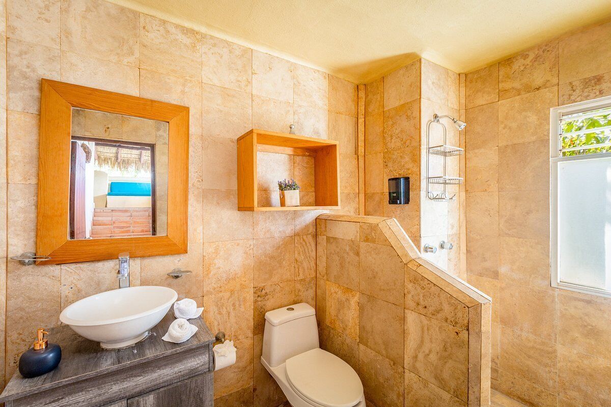Un baño con un lavamanos ovalado grande, inodoro, zona de ducha y estante de madera con artículos de aseo.