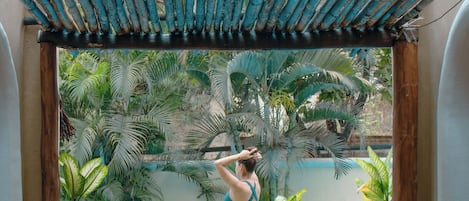 A woman in a swimsuit standing at the edge of a pool, surrounded by tropical plants and a wooden structure overhead.