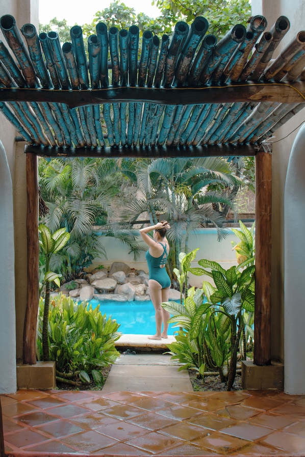 A woman in a swimsuit standing at the edge of a pool, surrounded by tropical plants and a wooden structure overhead.