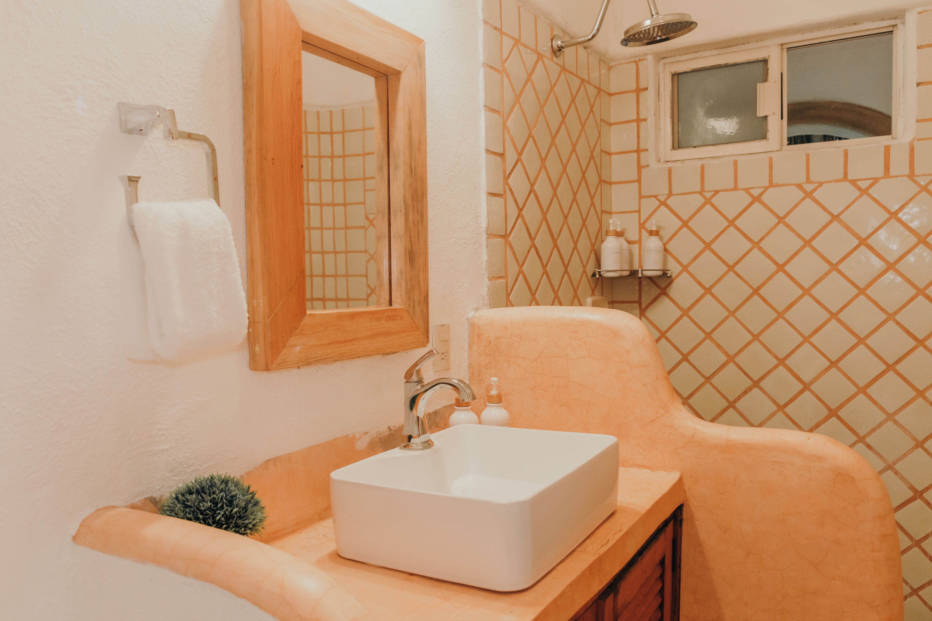 Un baño con un lavabo blanco, un espejo con marco de madera, una ducha con ventana y una pared con azulejos.