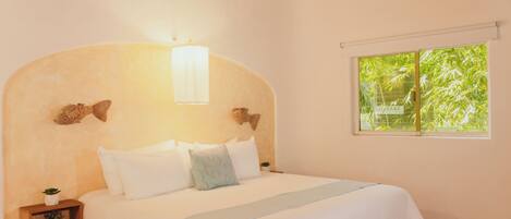 A bedroom with a bed, a nightstand with a plant, a window with a view of greenery, and a wall-mounted light fixture.
