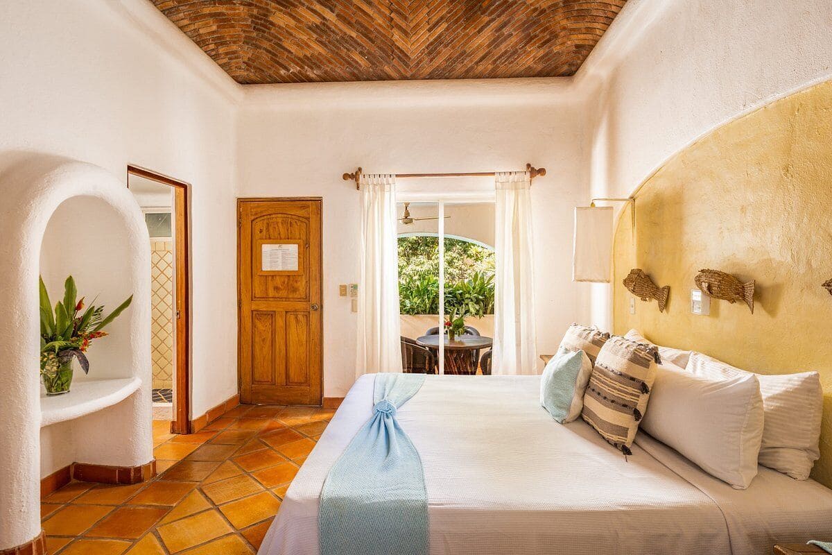 Une chambre d’hôtel avec un lit, une porte en bois, une fenêtre avec des rideaux, une petite table avec des chaises et une plante en pot.