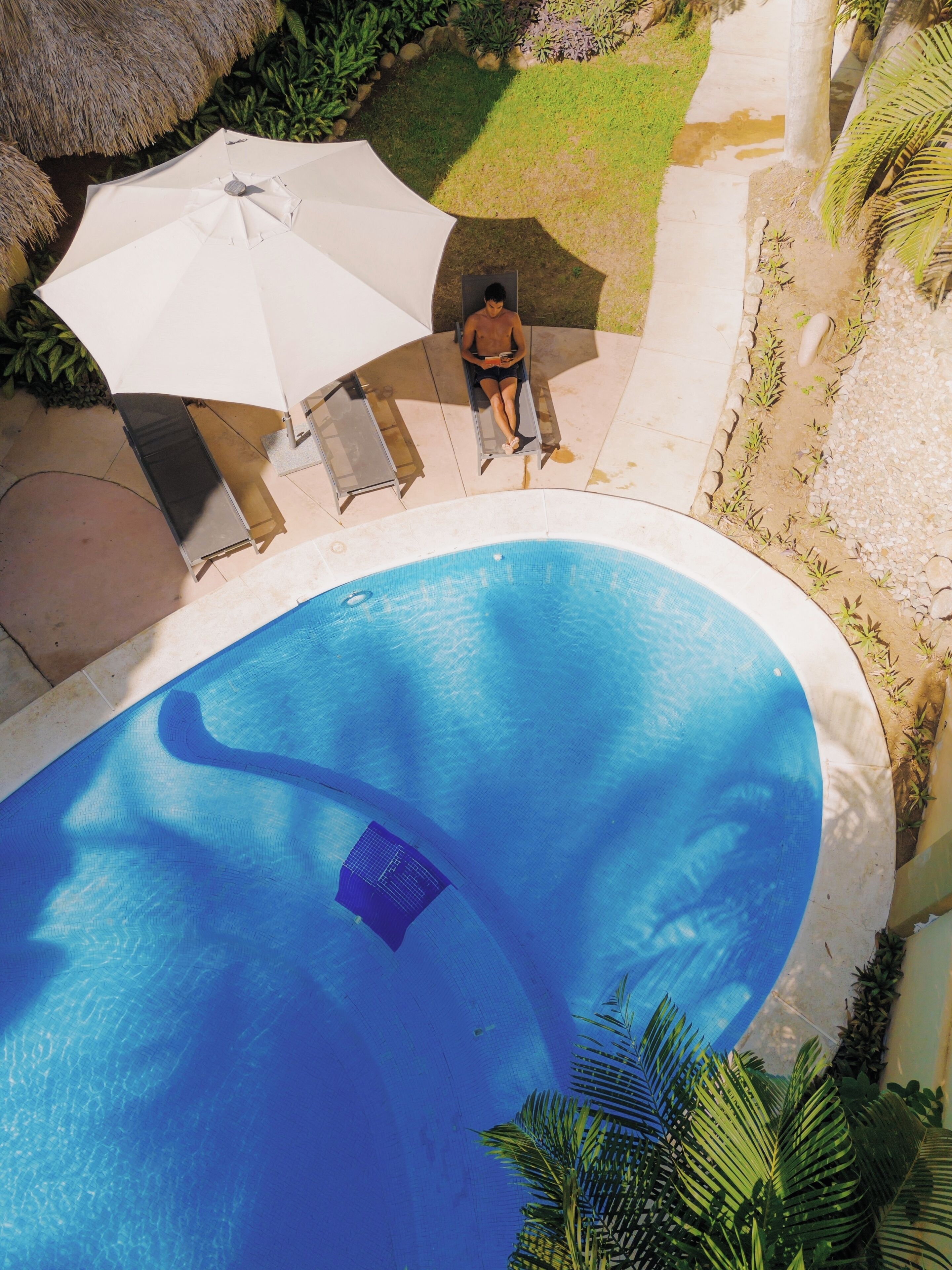 Une piscine avec une personne assise sur une chaise longue, à l’ombre d’un parasol blanc.