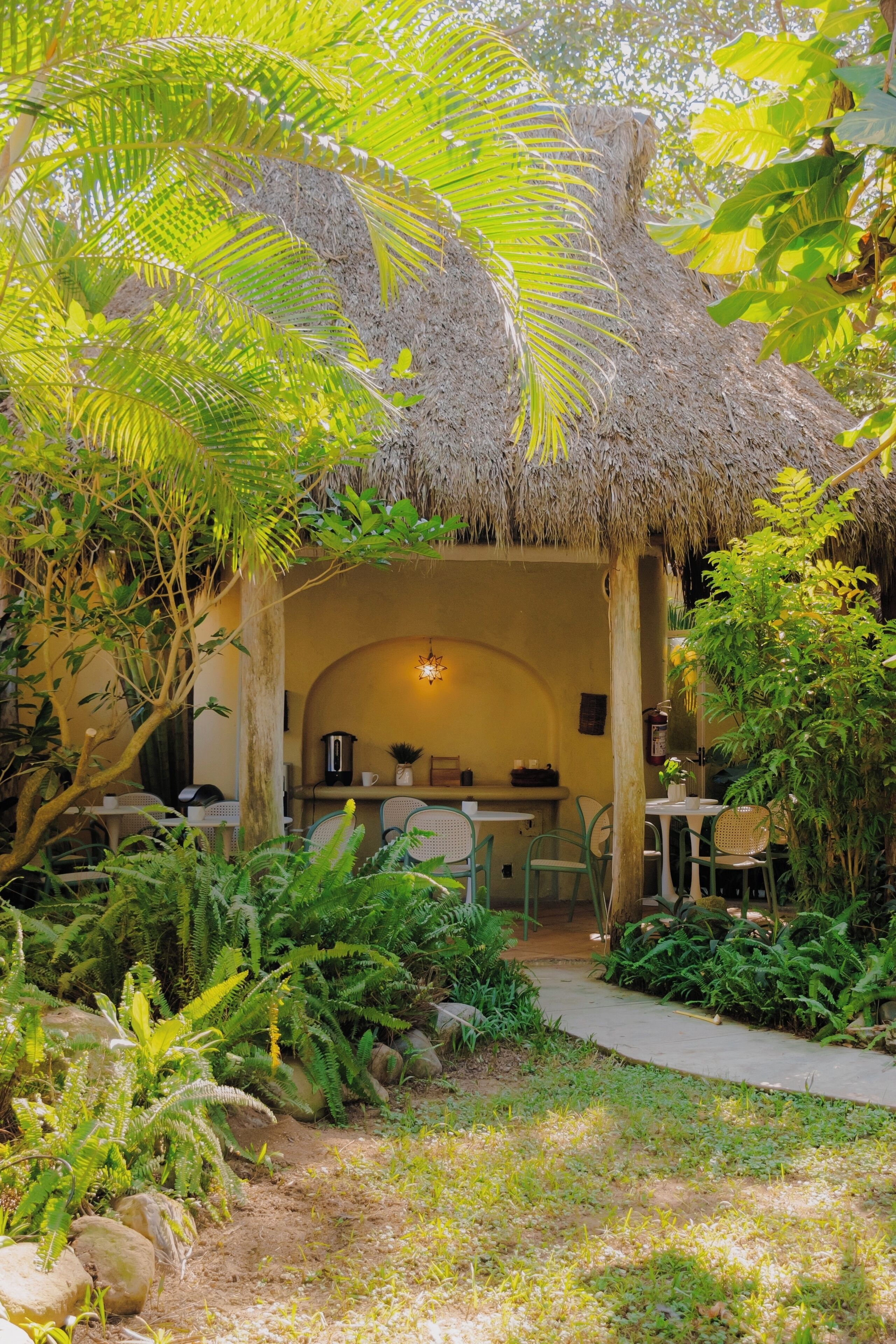 An outdoor dining area with thatched roof, surrounded by lush greenery and a pathway leading to a seating area.