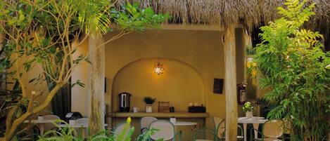 An outdoor dining area with thatched roof, surrounded by lush greenery and a pathway leading to a seating area.